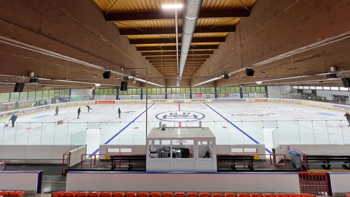 Eishalle Wiehl mit Eisfläche, Banden und Tribüne. Einige Eisläufer sind auf dem Eis zu sehen.