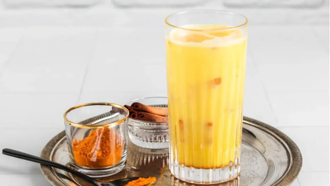 Ein Glas goldene Milch mit Kurkumapulver und Zimtstangen daneben.