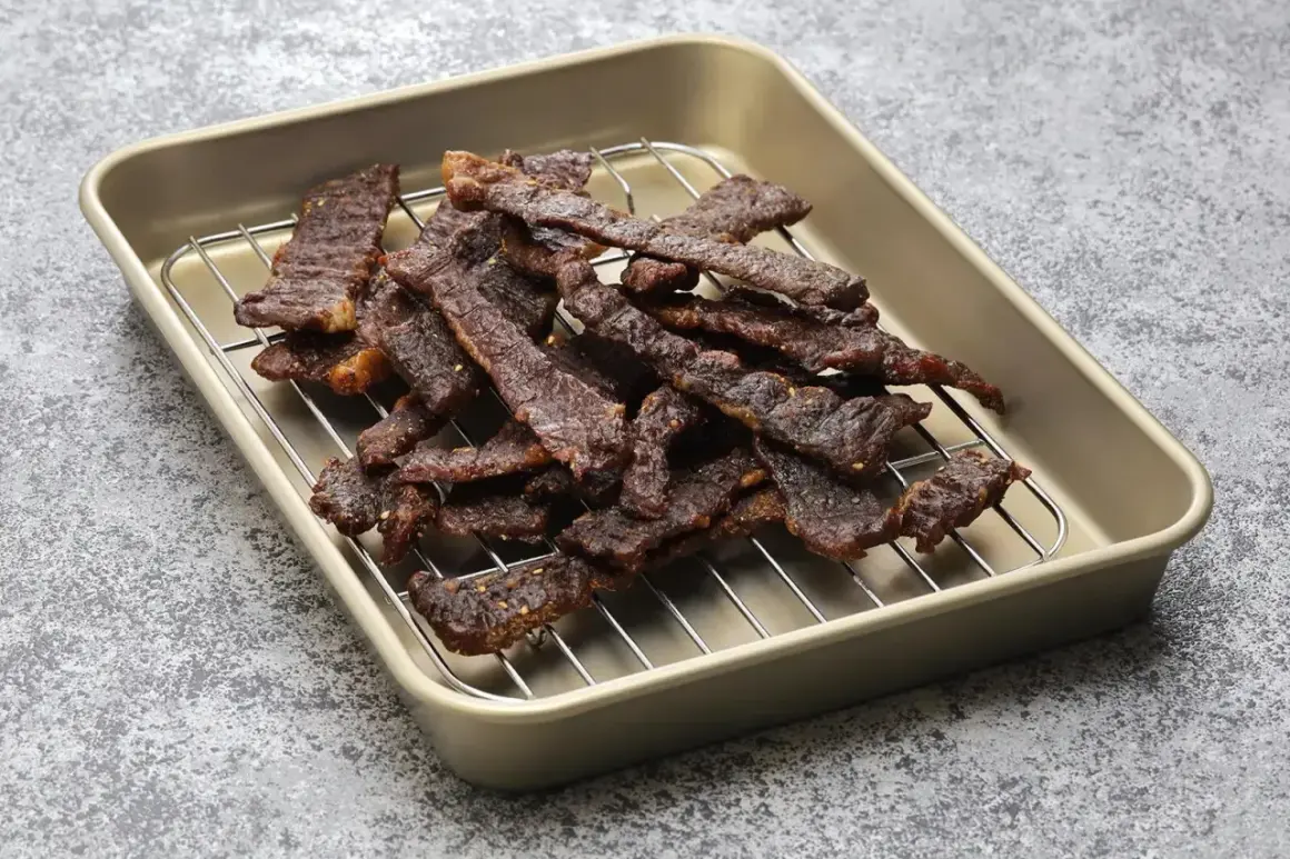 Stos cienkich, ciemnych plastrów wołowiny na metalowej kratce, gotowych do spożycia. Widać przyprawy, które nadają im smak, jak w klasycznej beef jerky marynata.