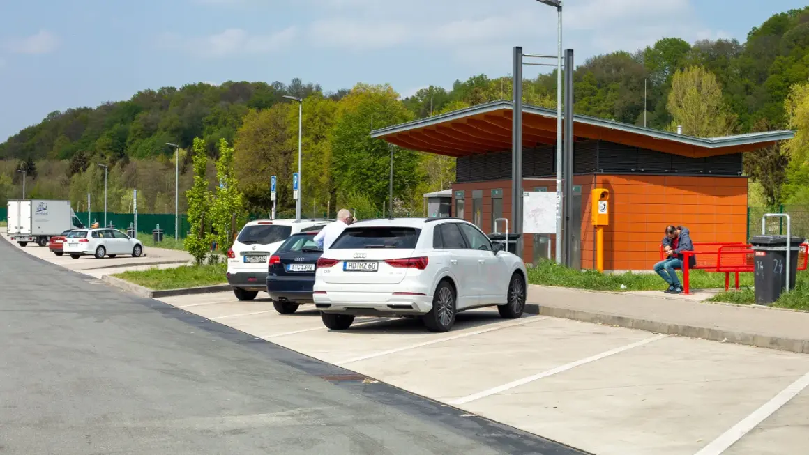 Stationnement sur une aire d'autoroute avec plusieurs voitures garées. Un homme est assis sur un banc rouge devant un bâtiment orange.