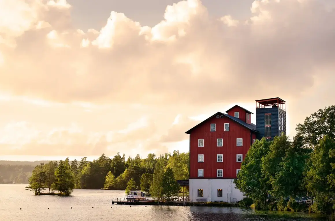 Skålleruds gård vid vattnet, med en röd byggnad och ett torn, omgiven av träd under en molnig himmel.