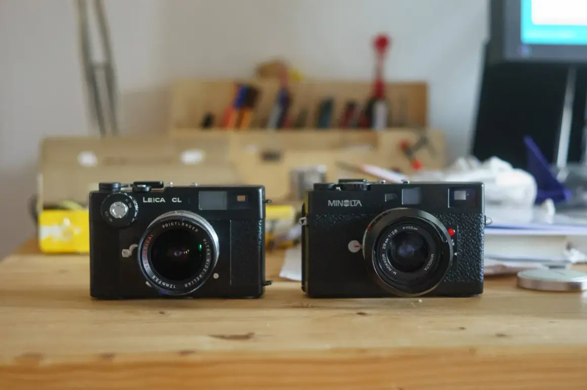 Two classic rangefinder cameras, a Leica CL and a Minolta, sit side-by-side on a wooden desk, ready for their next adventure.