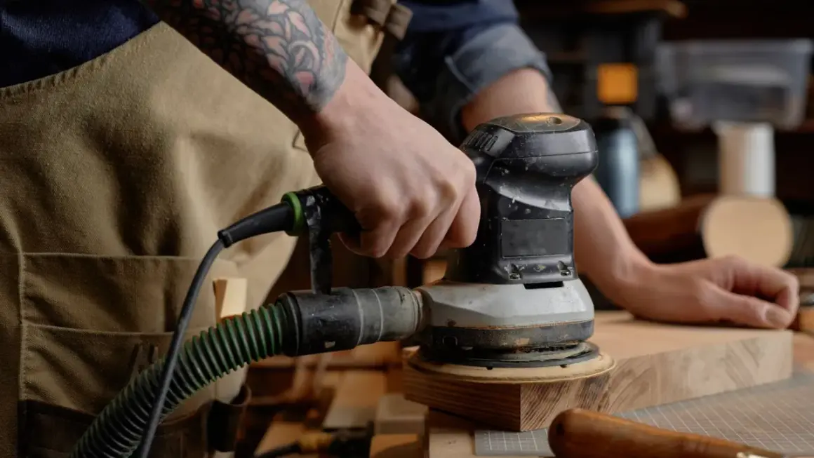 Main d'un artisan utilisant un outil pour poncer du bois. L'outil est relié à un aspirateur par un tuyau vert.