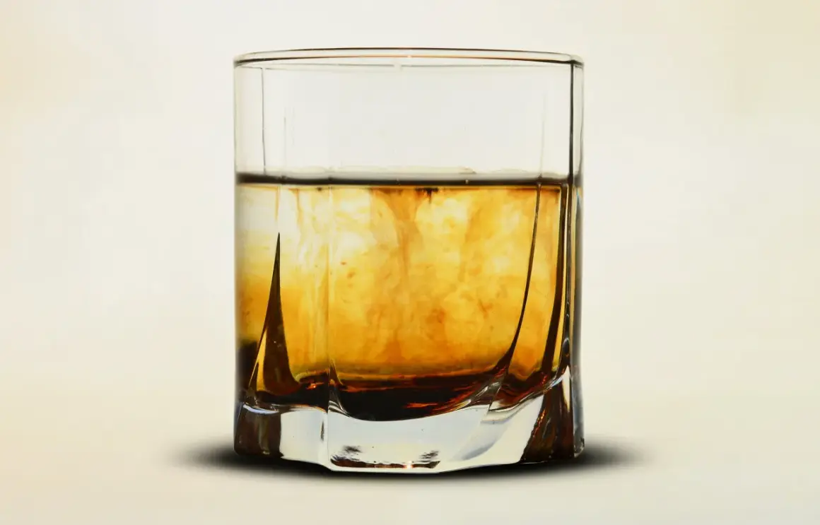 Szklanka z bursztynowym płynem, w którym rozpuszcza się ciemniejsza substancja, przypominająca proces, jak zrobic whiskey.