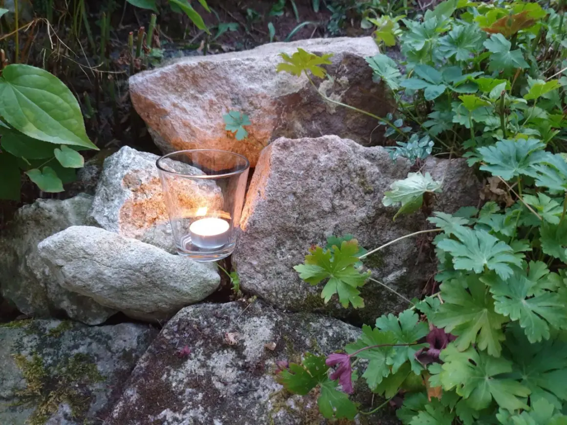 Ein Teelicht in einem Glas leuchtet zwischen Steinen und grünen Pflanzen. Ein Symbol für Hoffnung und einen bibelvers mut.