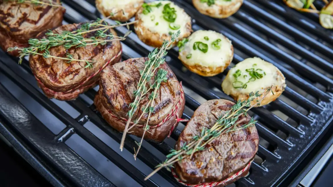 Saftige Rindersteaks mit Kräutern auf dem Holzkohlegrill. Daneben liegen gefüllte Champignons.