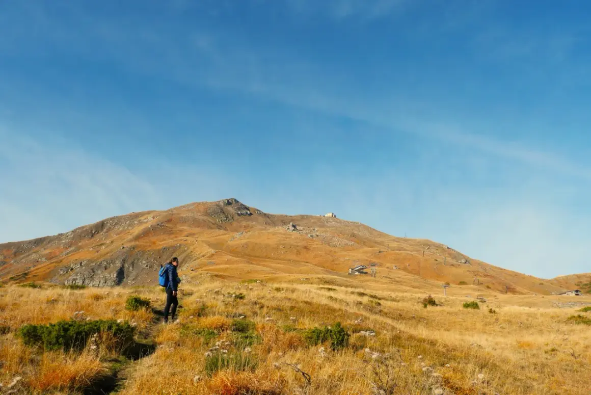 Escursionista in cammino verso il monte più alto dell'Emilia Romagna, con un paesaggio dorato e un cielo azzurro.