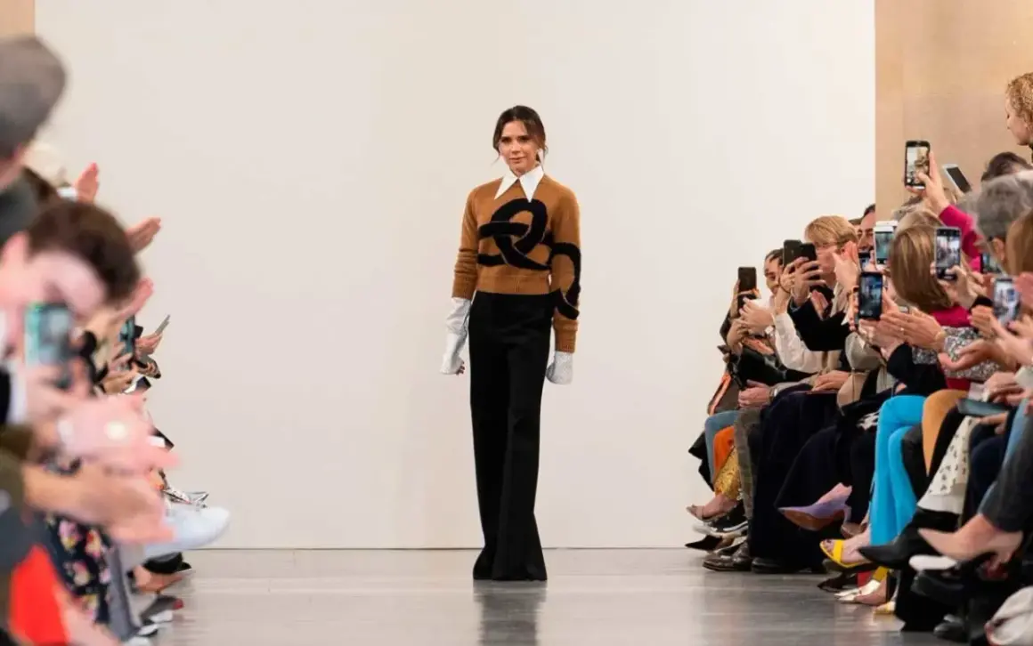 Victoria Beckham går på catwalken, klädd i en brun tröja med ett svart mönster och vida svarta byxor. Publiken, inklusive Harper Beckham, fotograferar med sina mobiler.