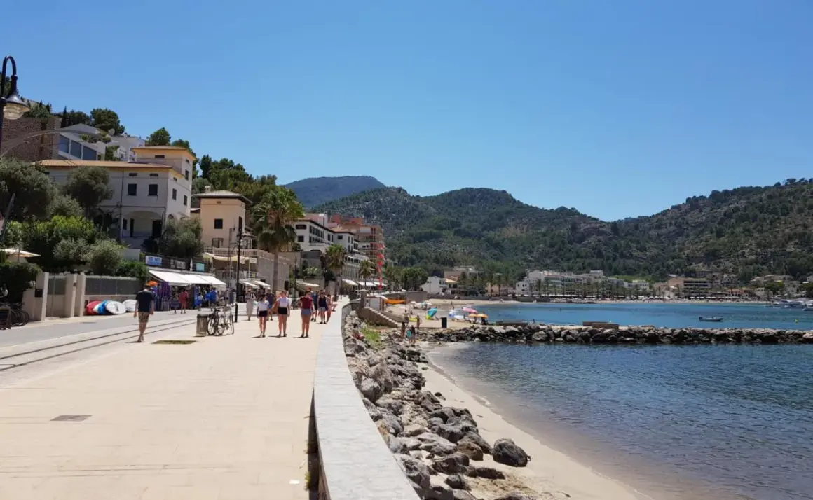 Promenad längs stranden i Port de Sóller med folk som njuter av solen och havet, omgivet av gröna berg.