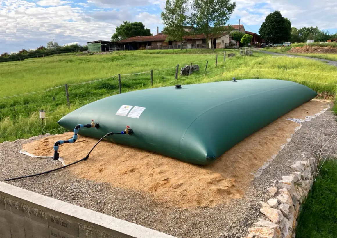 Gran depósito de agua de 20 litros, verde, instalado en un campo con una granja al fondo.