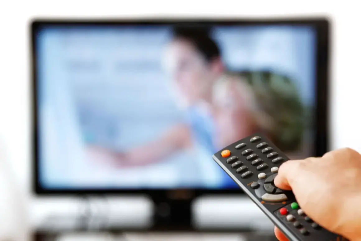 Dłoń trzymająca pilot do telewizora przed ekranem.