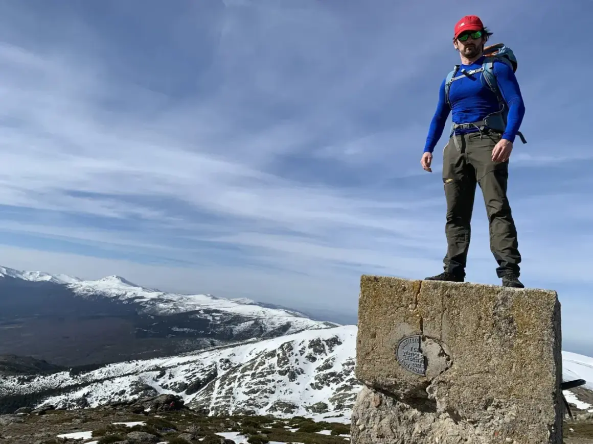 Hombre con mochila en la cima de un pico nevero, con vistas a montañas nevadas bajo un cielo azul con nubes.
