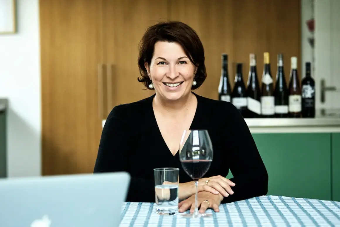 Eine lächelnde Frau, die wie ein **master of wine** wirkt, sitzt an einem Tisch mit Wein und Wasser. Im Hintergrund stehen mehrere Weinflaschen.
