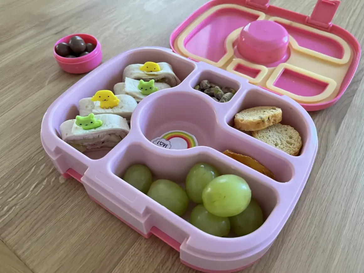 Różowa śniadaniówka do szkoły z przegródkami zawiera zawinięte tortille z uroczymi ozdobami, winogrona, orzeszki pistacjowe, krakersy i suszone mango.