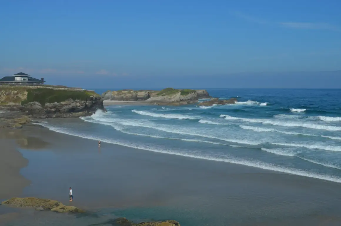 La **playa Os Castros** se extiende bajo un cielo azul, con olas rompiendo en la arena dorada. Una casa moderna corona el acantilado.