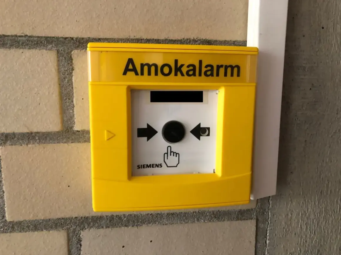 Gelber Amokalarm-Knopf an einer Ziegelwand. Ein Symbol zeigt eine Hand, die auf den Knopf drückt, um einen Amoklauf an einer Schule zu melden.