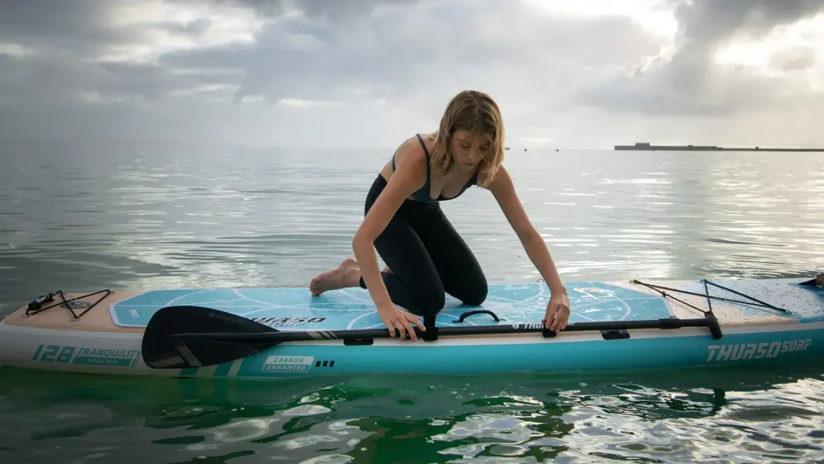 Donna in ginocchio su un sup, pronta a pagaiare. La tavola galleggia su acqua calma sotto un cielo nuvoloso.