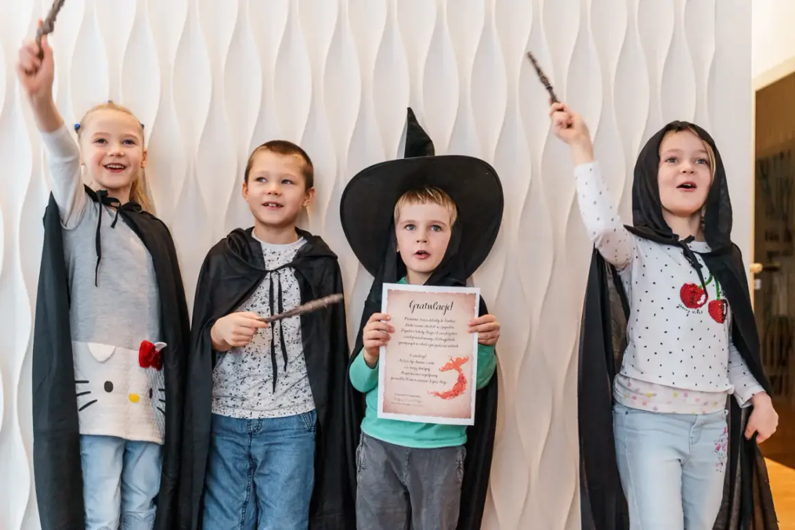 Cztery dzieci w strojach czarodziejów z różdżkami. Jeden chłopiec w kapeluszu czarownicy trzyma dyplom.