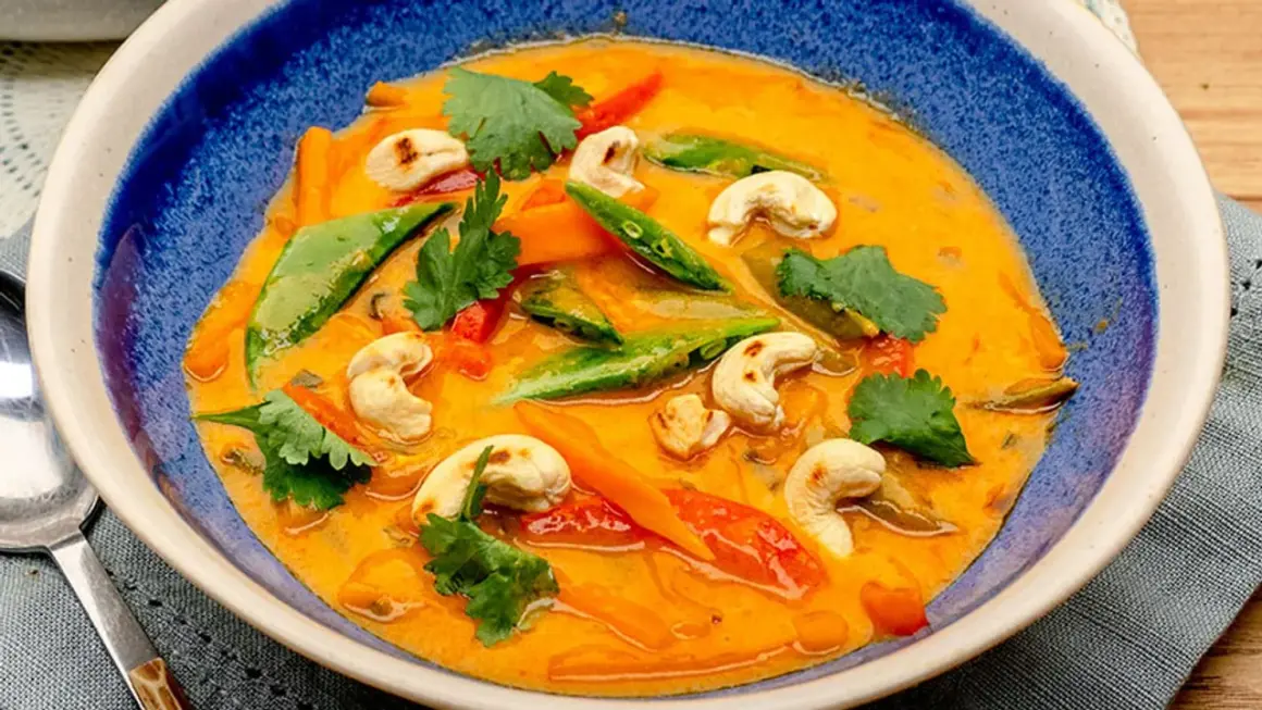 Ein cremiges rotes Thai Curry mit Cashewkernen, Zuckerschoten und Karotten in einer blauen Schüssel.