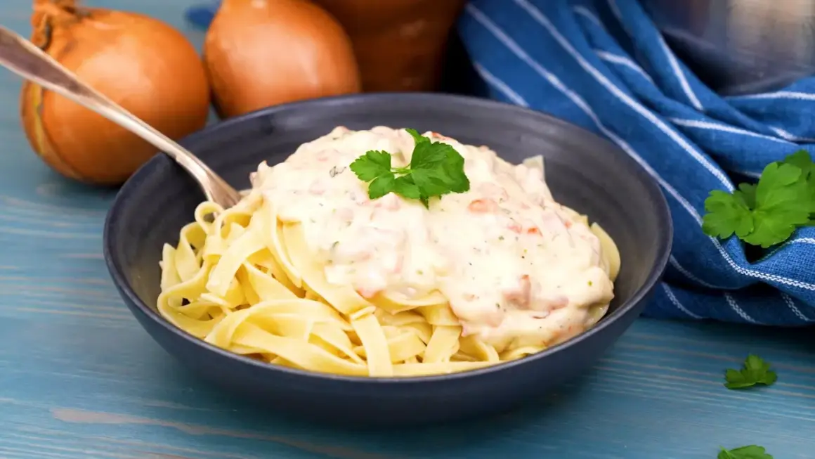 En skål med tagliatelle toppad med krämig ost och skinksås, garnerad med persilja. Två gula lökar syns i bakgrunden.