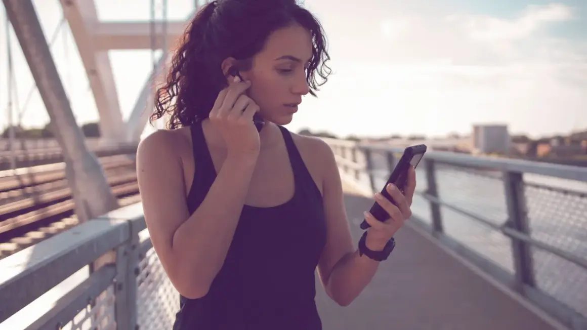 Frau auf einer Brücke, die sich Kopfhörer einsetzt und auf ihr Handy schaut. Sie ist bereit für ihre täglichen 10.000 Schritte.