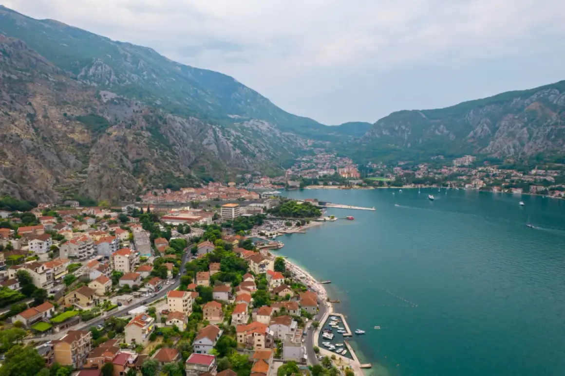 Widok na zatokę i miasto Kotor w Czarnogórze. Domy z czerwonymi dachami schodzą ku turkusowej wodzie, a nad wszystkim górują strome, skaliste wzgórza.