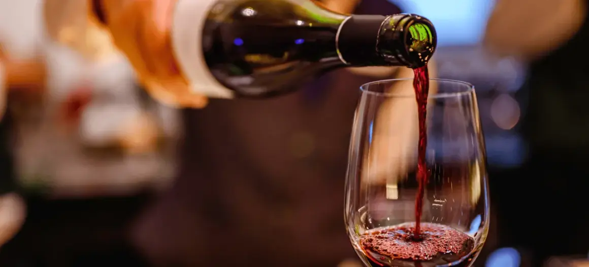 Czerwone wino leje się z butelki do kieliszka, tworząc piankę. Widać bogactwo **tannin w winie**, które nadają mu głębię.
