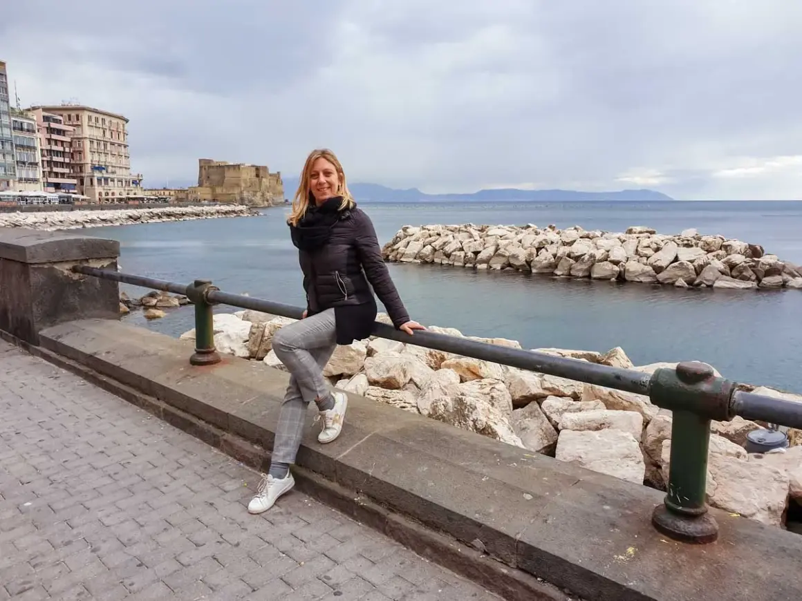 Donna sorridente sul lungomare di Napoli, con Castel dell'Ovo e il mare sullo sfondo. Un'ottima idea per cosa vedere a Napoli centro.