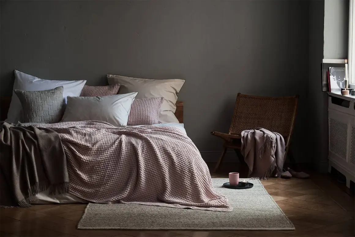 Gemütliches Schlafzimmer mit Bett in altrosa und grau, Korb-Sessel und Kerzenlicht.
