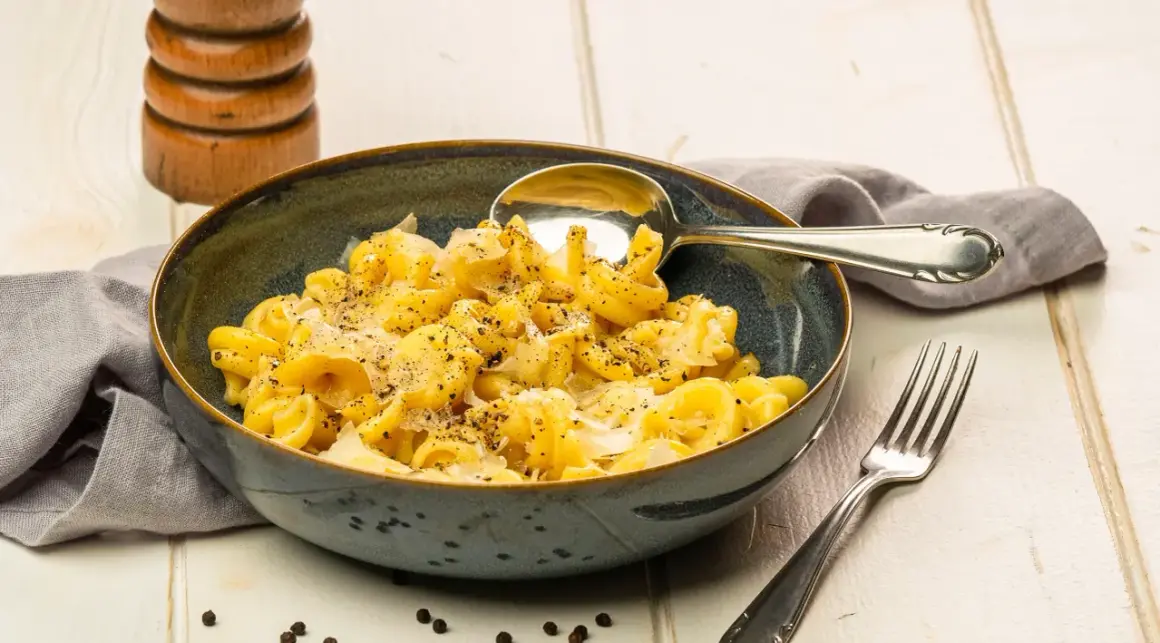 Eine Schüssel Pasta mit cremiger cacio e pepe sauce, bestreut mit frisch gemahlenem Pfeffer. Daneben liegen ein Löffel und eine Gabel.