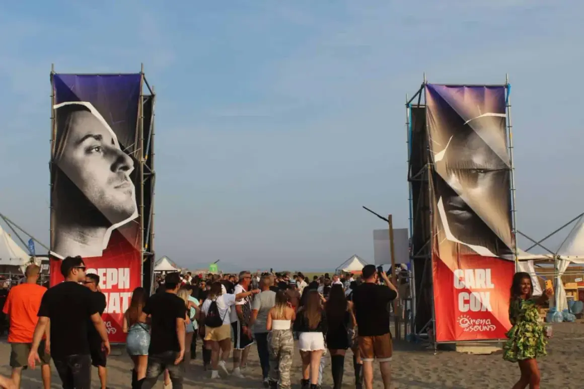 Ingresso per il festival con gigantografie di Carl Cox e Joseph Capriati. La folla si muove sulla sabbia.