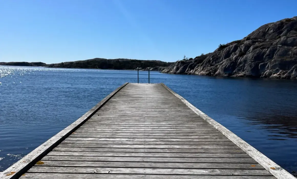 Träbrygga leder ut mot det glittrande vattnet vid Grebbestad strand. Klippor och öar i bakgrunden under en klarblå himmel.