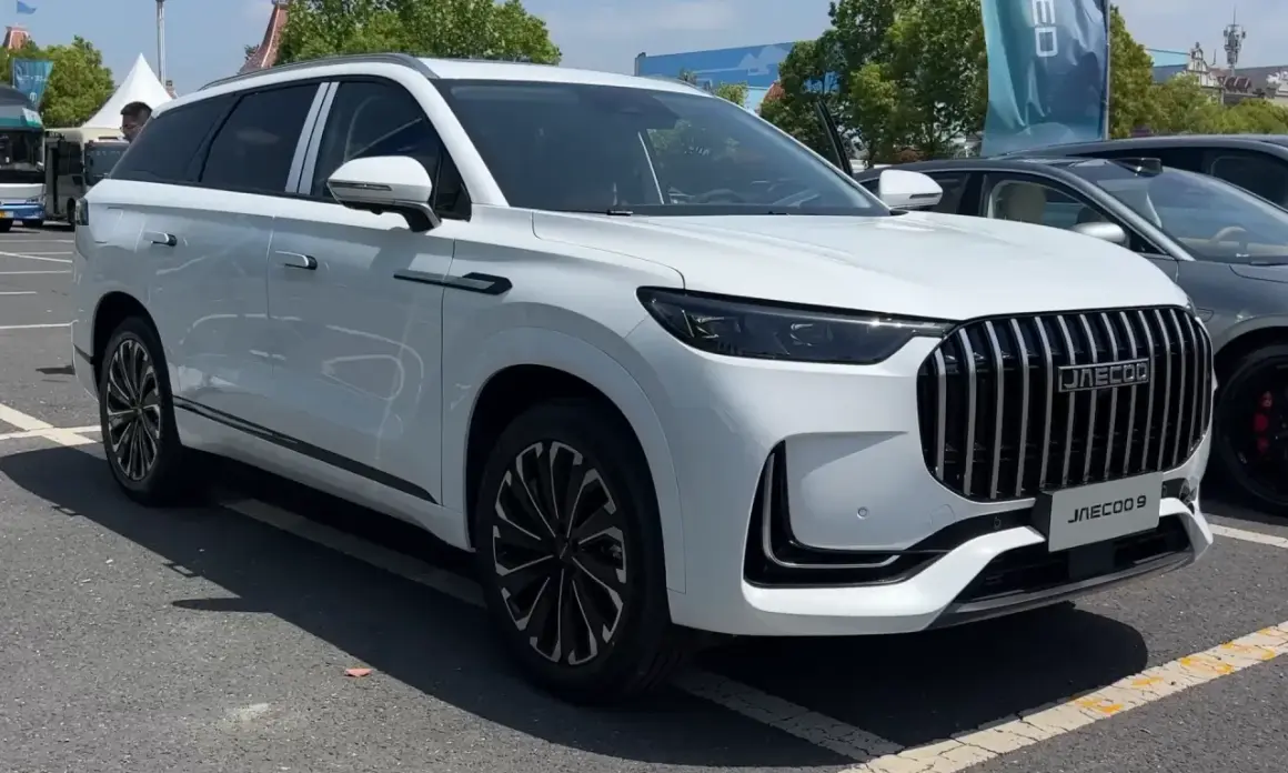 Biały SUV Jaecoo J9 z chromowanym grillem i czarnymi felgami stoi na parkingu.