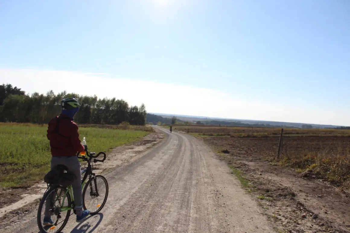 Rowerzysta na szutrowej drodze, która wydaje się być częścią najdłuższej trasy rowerowej w Polsce. Słońce świeci na błękitnym niebie.