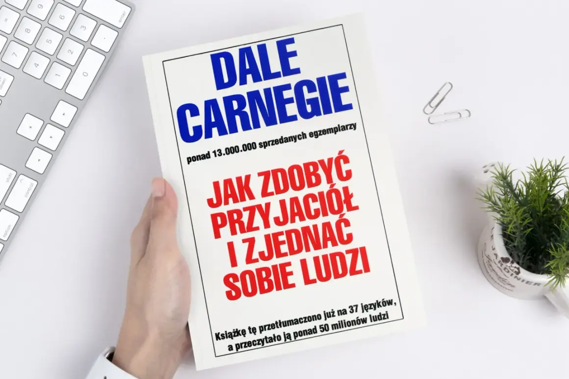 Książka "Jak zdobyć przyjaciół i zjednać sobie ludzi" autorstwa Dale'a Carnegie, trzymana w dłoni, leży obok klawiatury i rośliny.