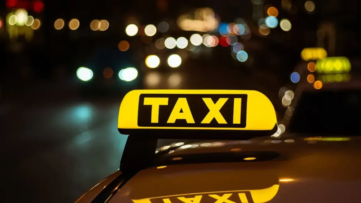 Żółty znak TAXI na dachu samochodu nocą. W tle rozmyte światła miasta, sugerujące licencję taxi w ruchu.