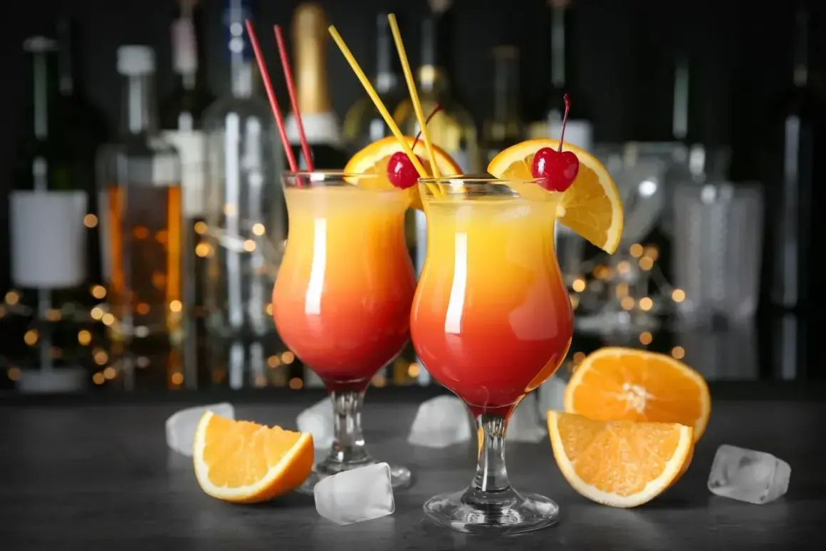 Dwa drinki Tequila Sunrise, ozdobione pomarańczą i wiśnią, stoją na barze. W tle widać butelki alkoholu i migoczące światełka. Idealne na wieczór z przyjaciółmi, jak w najlepszych lokalach Baczewski.