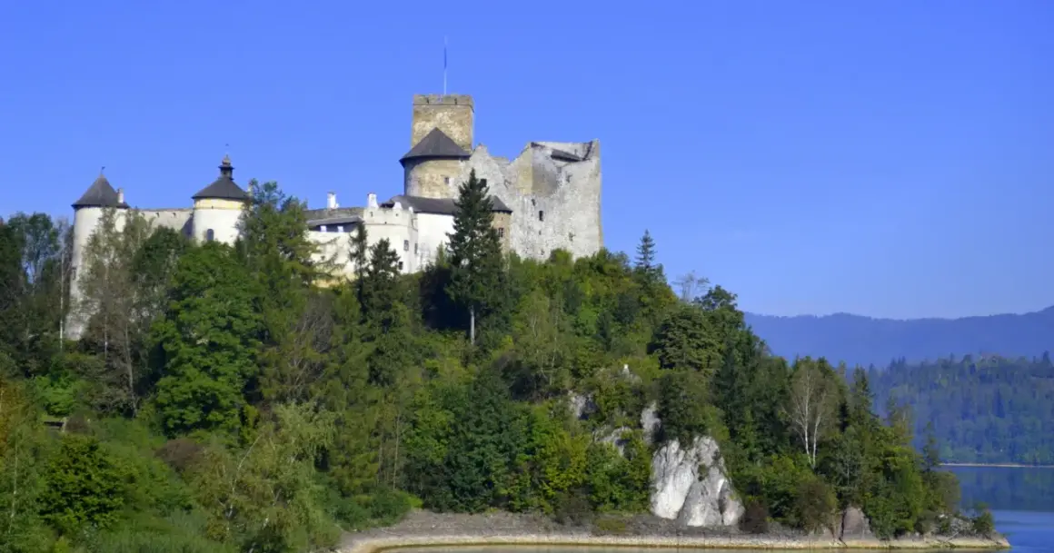 Zamek w Niedzicy, majestatyczna twierdza na wzgórzu, otoczona zielenią, z widokiem na jezioro i góry.