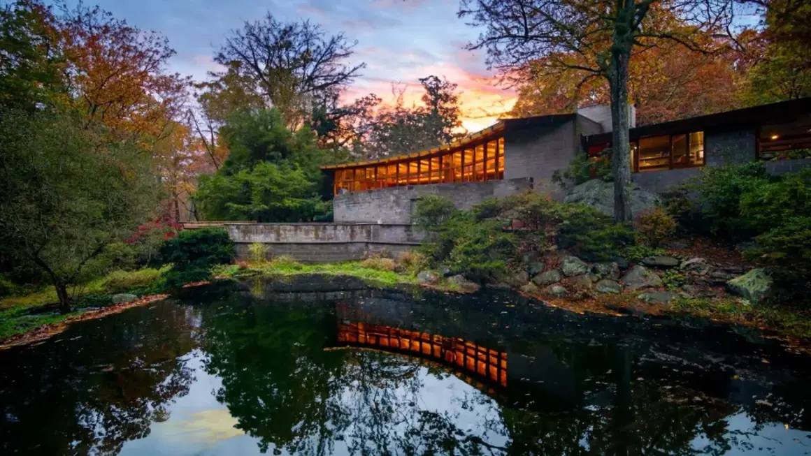 The last Wright house, a modern masterpiece, reflects warmly in a tranquil pond amidst autumn foliage.