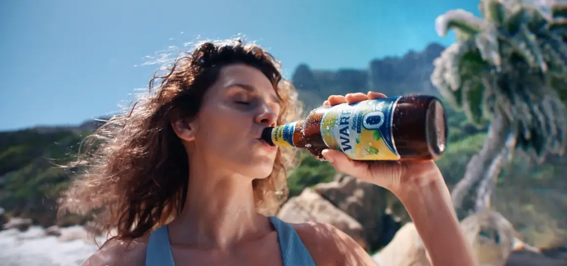 Kobieta pije piwo Warka Radler 0,0% na plaży. Idealne na upalne dni, gdy chcesz orzeźwienia bez procentów.