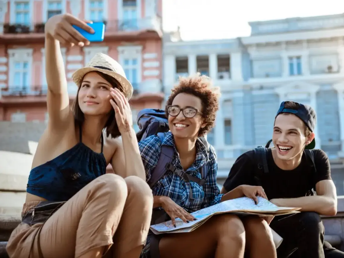 Tres amigos sonríen mientras se toman una selfie en la calle, planeando cómo conocer gente para viajar y explorar la ciudad.
