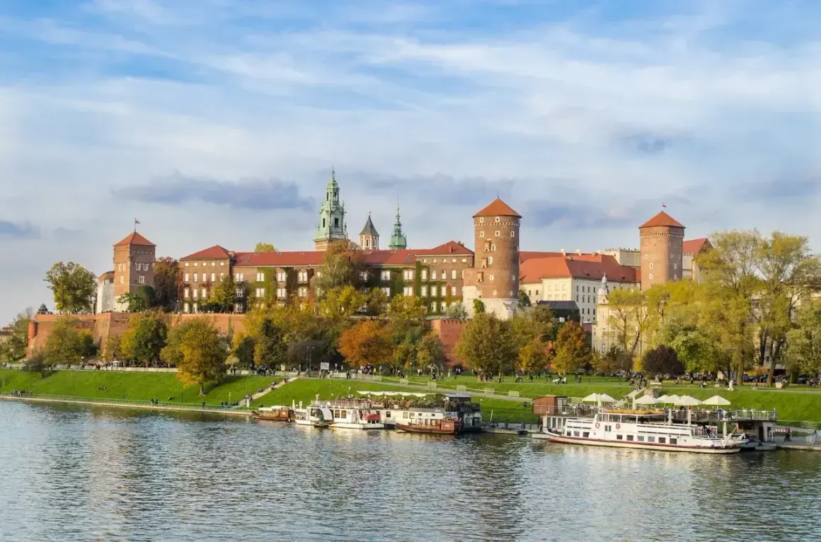 Malownicze miasto nad Wisłą z Wawelem w jesiennej odsłonie. Statki wycieczkowe cumują przy brzegu, a na trawniku odpoczywają spacerowicze.