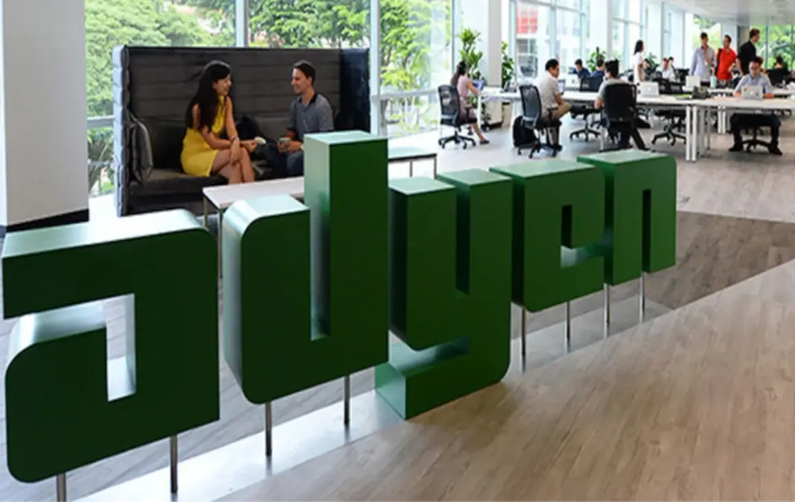 Zielone, przestrzenne litery z napisem "adyen" w nowoczesnym biurze. W tle ludzie pracują przy biurkach i rozmawiają.