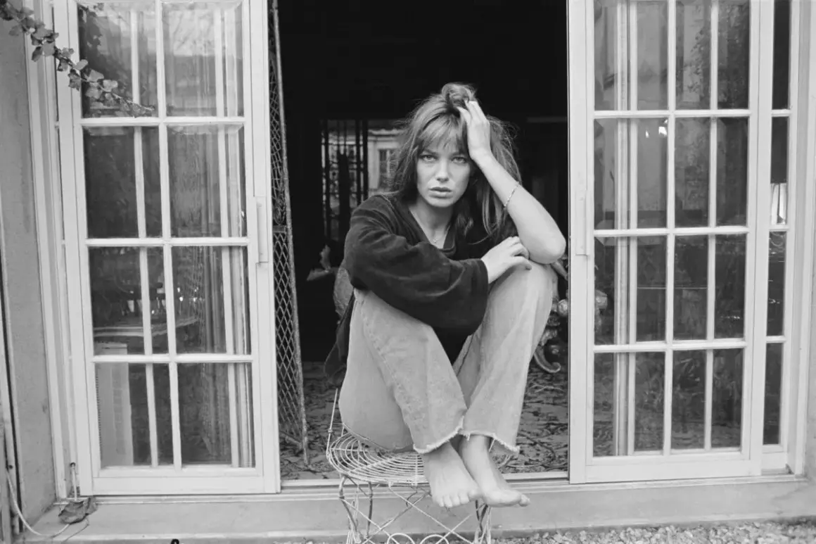 Jane Birkin mit typischen 70er Frisuren, lässig auf einem Stuhl sitzend, vor einer Tür mit Sprossenfenstern.
