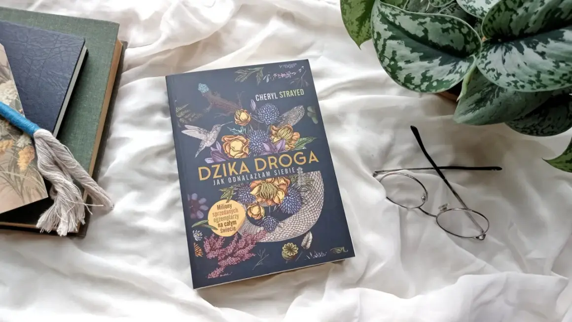 Okładka książki "Dzika droga" z motywami roślinnymi i zwierzęcymi, obok okulary i inne książki.