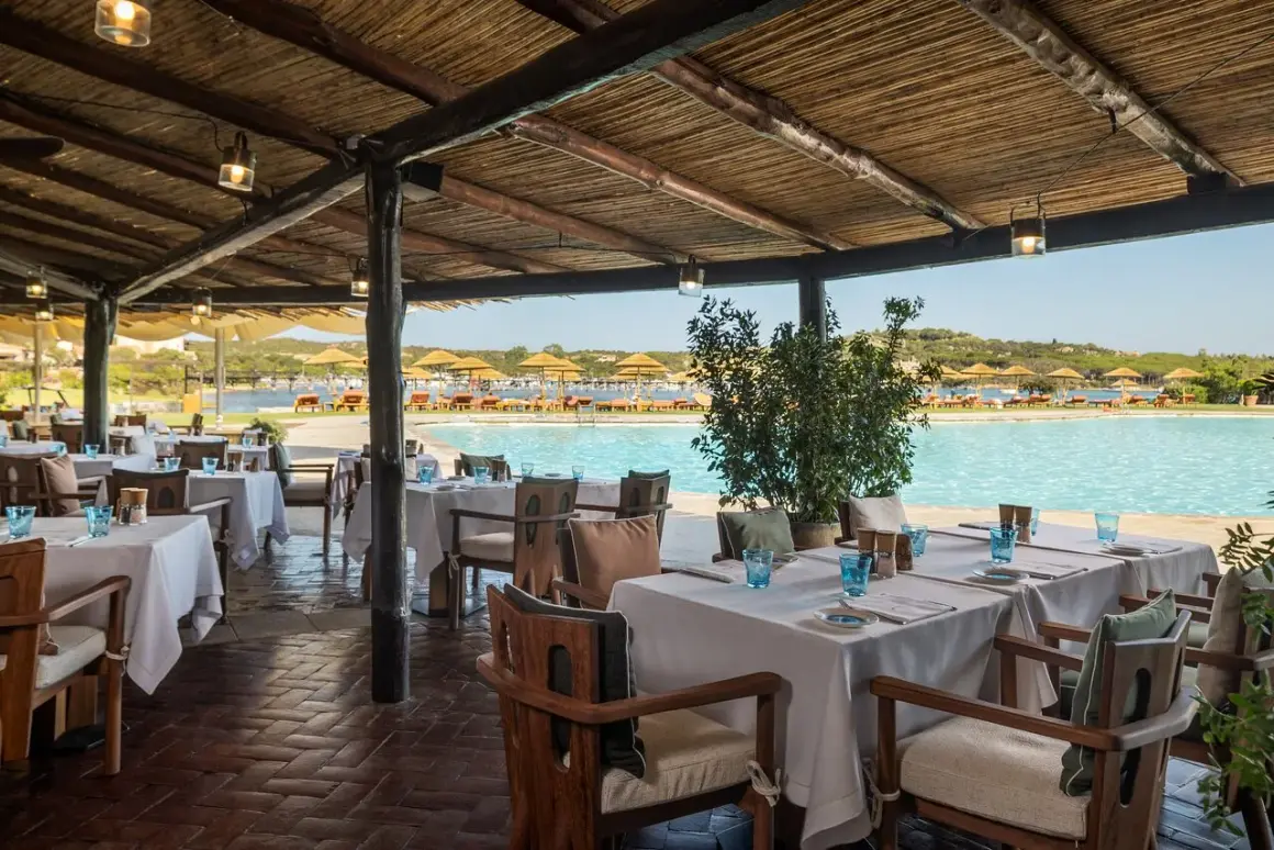 Tavoli apparecchiati sotto un tetto di paglia, con vista sulla piscina e sulla spiaggia del **hotel Cala di Volpe**.
