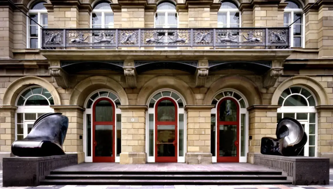 Vor dem Museum Wuppertal stehen zwei abstrakte Skulpturen. Die Fassade des Gebäudes ist aus hellem Stein, mit roten Türen und verzierten Balkonen.