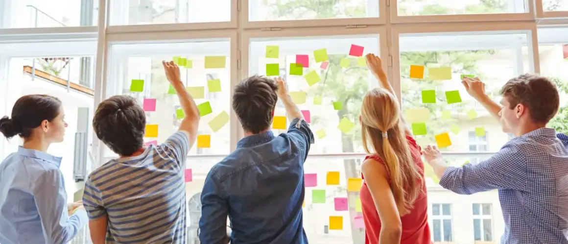 Fünf Personen planen an einem Fenster mit bunten Haftnotizen. Sie arbeiten an ihrer kaufmännischen Ausbildung nebenberuflich.