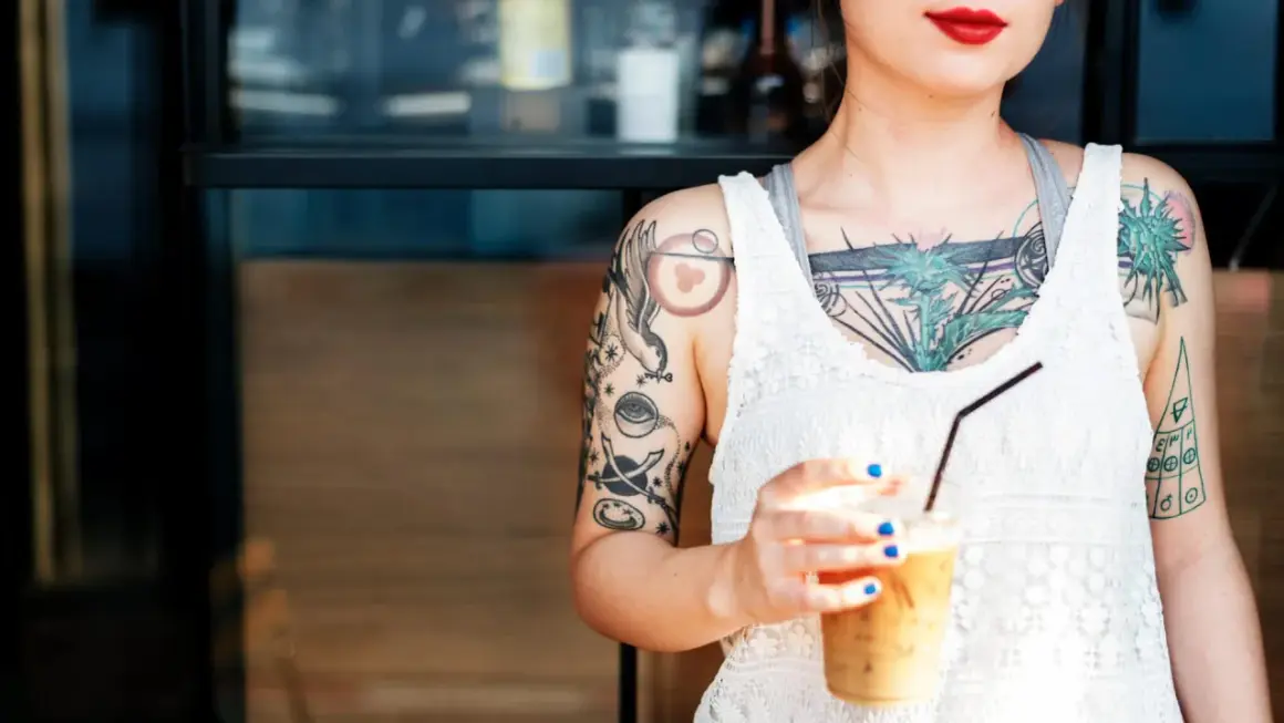 Junge Frau mit vielen Tattoos, darunter ein Vogel und ein Auge. Sie hält einen Eiskaffee. Ein Tattoo ab 16, das ihre Persönlichkeit unterstreicht.