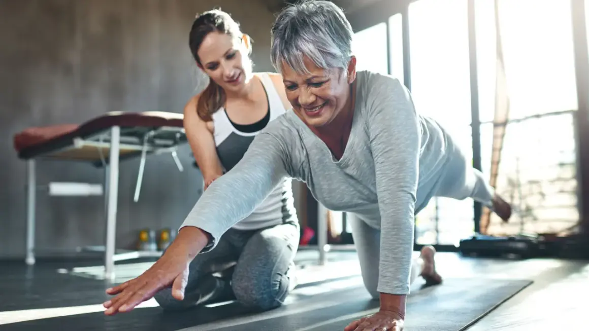 Une femme âgée sourit en faisant un exercice de renforcement avec l'aide d'une coach. Le sport est essentiel pour la rééducation après un cancer du sein.